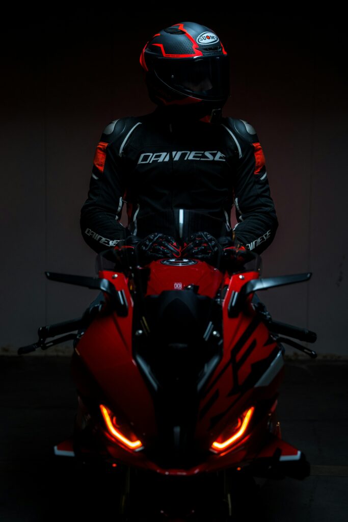 A motorcyclist in black protective gear sits on a red sports bike under dim lighting.